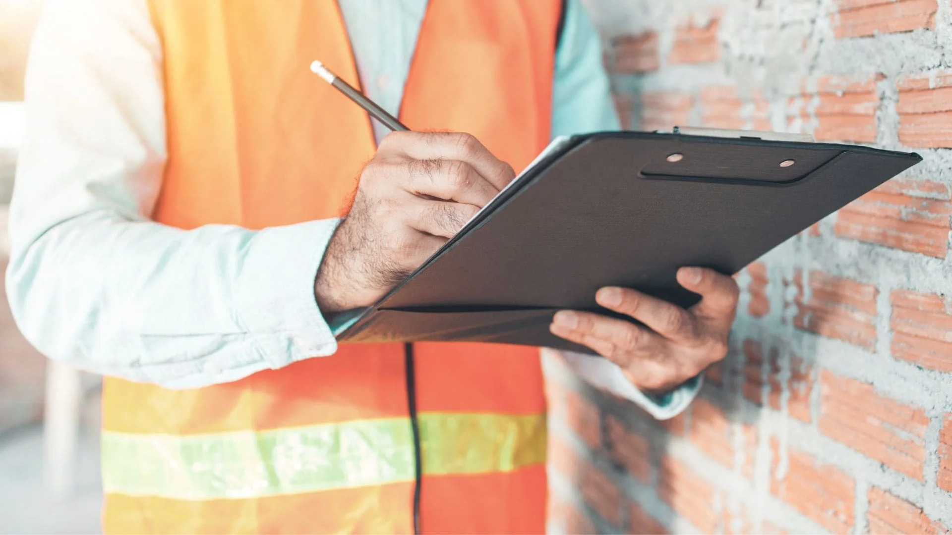 A contractor in a hi-vis vest taking notes on a clipboard beside a brick wall.