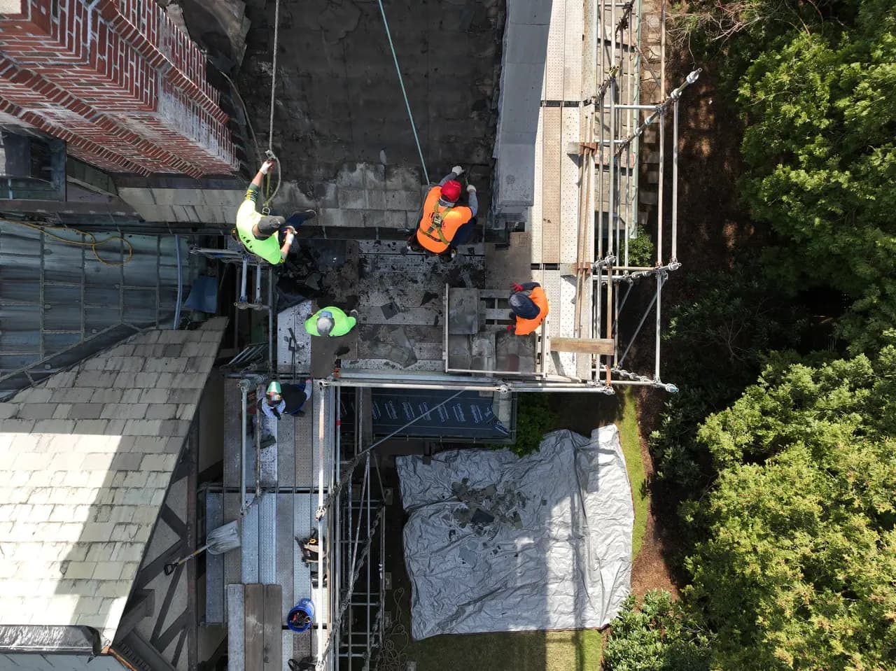 Top-down view of a Crown crew on a steep slate roof, with scaffolding and a brick chimney.